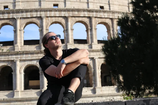 Young male caucasian tourist sitting near Colosseum background in Rome, Italy.