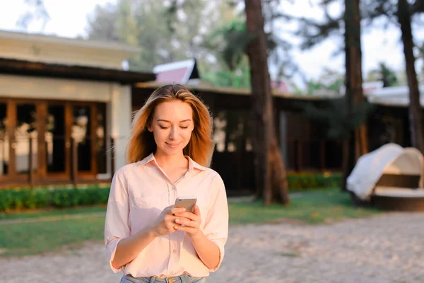 Mujer joven charlando por smarptone cerca de la casa del resort, usando blusa y pantalones cortos . — Foto de Stock