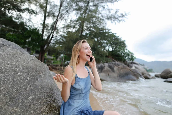 Kum deniz ve taş yakın oturan, smartphone ile konuşmak ve gösterilen genç sarışın kız yaşasın.