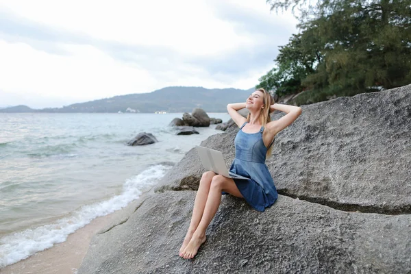 Deniz kıyısında laptop ile çalışma ve taş üzerine oturan genç beyaz kadın.
