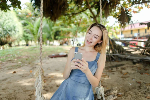 Salıncak sürme ve smatrphone, kum ve ağaç arka planda kullanarak mutlu kadın.
