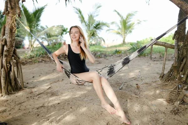 Junges Mädchen sitzt auf Hängematte mit Palmen und Sand im Hintergrund und trägt schwarzen Badeanzug. — Stockfoto