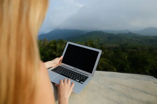 Chica joven que trabaja con el ordenador portátil en el fondo de las montañas verdes, con camisa gris . — Foto de Stock