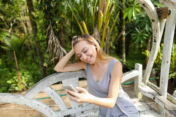 Avuç içinde geçmiş, salıncakta oturan Smartphone kullanan genç mutlu kız. Modern teknoloji ve egzotik tatil dinlenme kavramı.