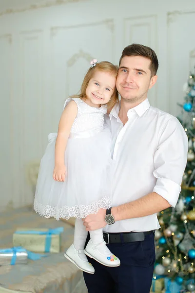 Νέος πατέρας κρατώντας την μικρή κόρη του φορώντας λευκό φόρεμα κοντά στο χριστουγεννιάτικο δέντρο. — Φωτογραφία Αρχείου