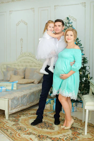Kocası ve Noel ağacı yanında küçük kızı ile ayakta mavi elbise giyen hamile Avrupa kadın. Kış tatili ve aile fotoğraf oturumu kavramı.
