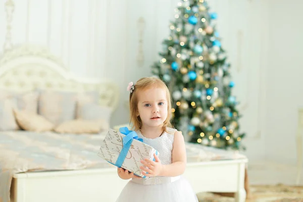 Kleines süßes Mädchen, das Geschenke hält, weißes Kleid trägt und im Schlafzimmer mit Weihnachtsbaum steht. — Stockfoto