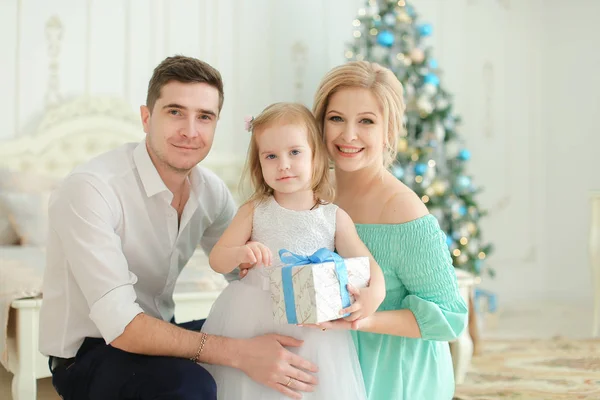 Sorrindo mulher e homem sentado com a filhinha mantendo presente, árvore de Natal no fundo . — Fotografia de Stock