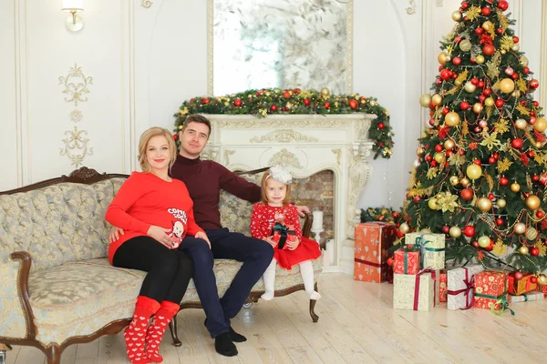 Mujer embarazada sentada con el hombre y la hija cerca del árbol de Navidad y chimenea decorada . — Foto de Stock