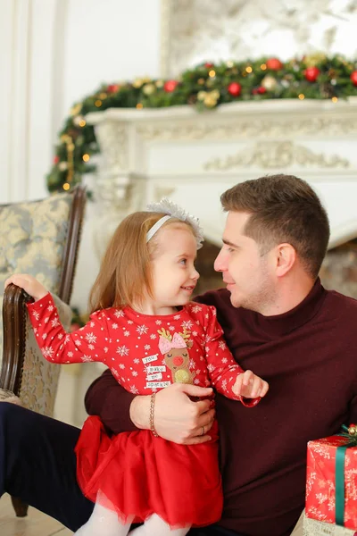 Padre europeo sentado con su hija pequeña cerca de la chimenea decorada y manteniendo regalos . — Foto de Stock