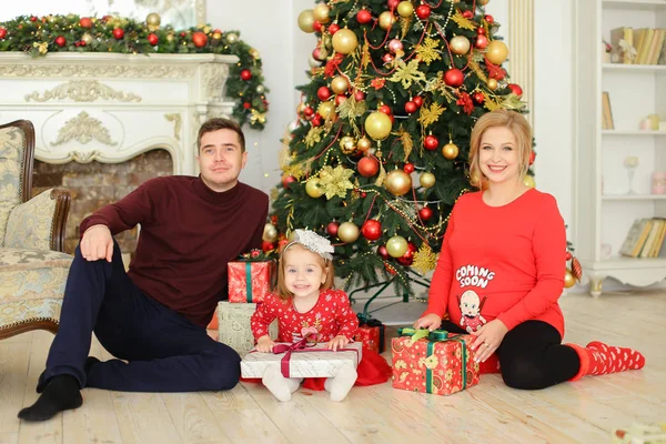 Kleine blonde Tochter sitzt mit Vater und schwangerer Mutter am Weihnachtsbaum und bewahrt Geschenke auf. — Stockfoto