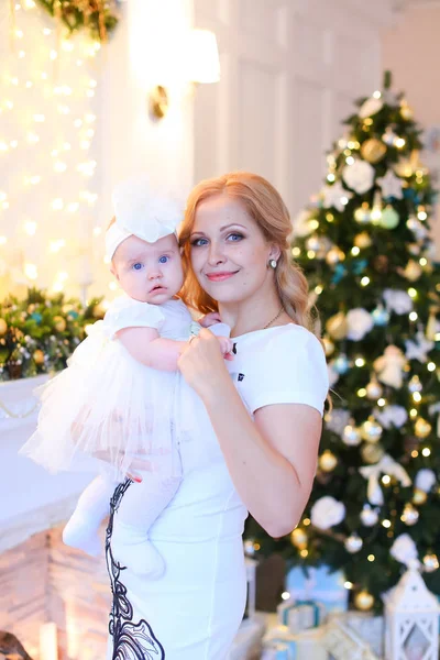 Jovem mãe sorridente vestindo vestido branco e mantendo pouco bebê feminino perto da árvore de Natal . — Fotografia de Stock