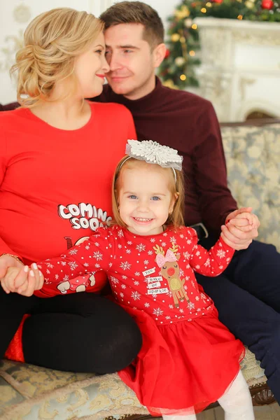 Niña sonriente sentada con el padre y la madre embarazada cerca de la chimenea decorada . — Foto de Stock