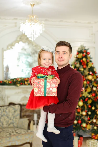 Junger kaukasischer Vater hält kleine Tochter mit Geschenk, Weihnachtsbaum im Hintergrund. — Stockfoto