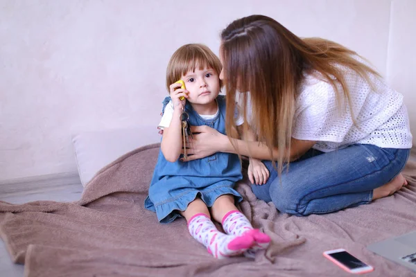 Giovane madre seduta vicino a piccola figlia e smartphone sul letto . — Foto Stock