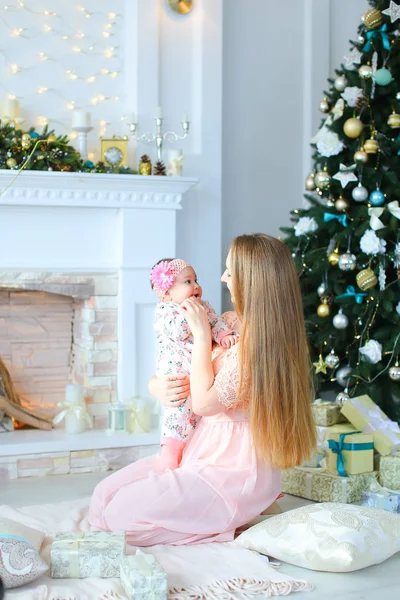 Amerikansk mor innehav dotter i armar nära inredda spis och julgran, klädd i rosa klänning. — Stockfoto