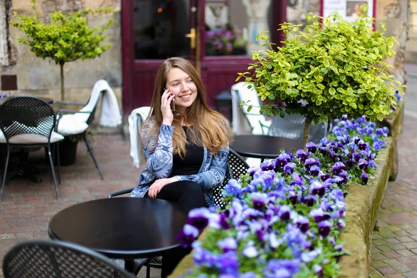 Young woman sitting at street cafe near blue flowers and green plants and talking by smartphone.