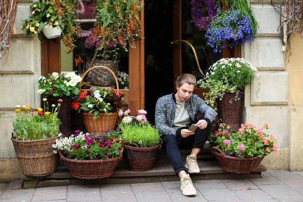 Young european man chatting by smartphone and sitting at flower shop near bouquets in baskets.