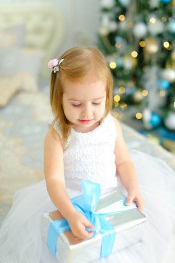 Küçük kız yatakta mevcut ve giyen beyaz elbise ile oturmuş dekore edilmiş arka planda köknar ağacı.