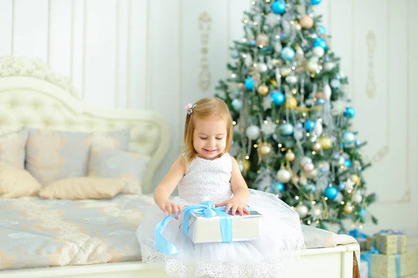 Kleines blondes kaukasisches Mädchen sitzt auf dem Bett mit Geschenk und trägt weißes Kleid, geschmückter Tannenbaum im Hintergrund. — Stockfoto