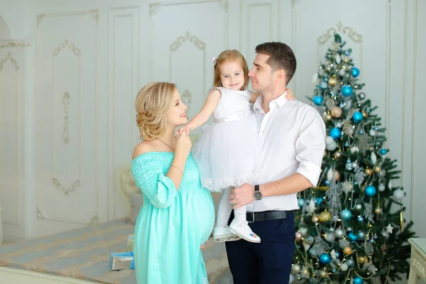 Embarazada joven mujer de pie cerca de marido mantener pequeña hija cerca de árbol de Navidad . — Foto de Stock