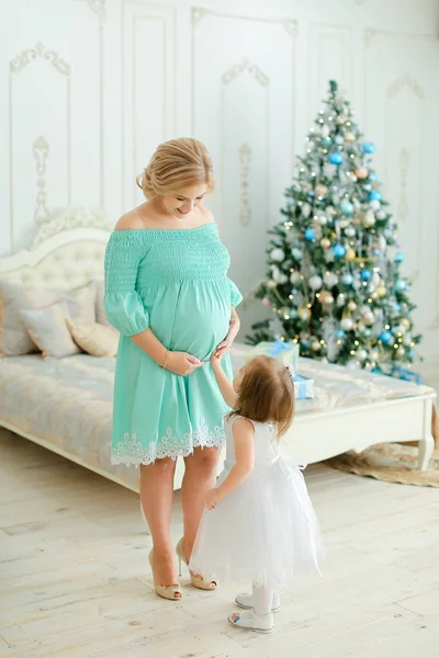 Schwangere kaukasische Frau trägt blaues Kleid, umarmt Bauch und steht mit kleiner Tochter neben Weihnachtsbaum im Schlafzimmer. — Stockfoto