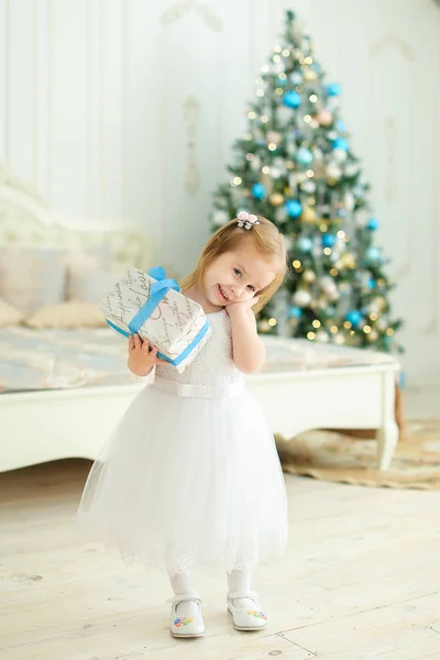Kleines glückliches Mädchen hält Geschenk, trägt weißes Kleid und steht im Schlafzimmer mit Weihnachtsbaum. — Stockfoto
