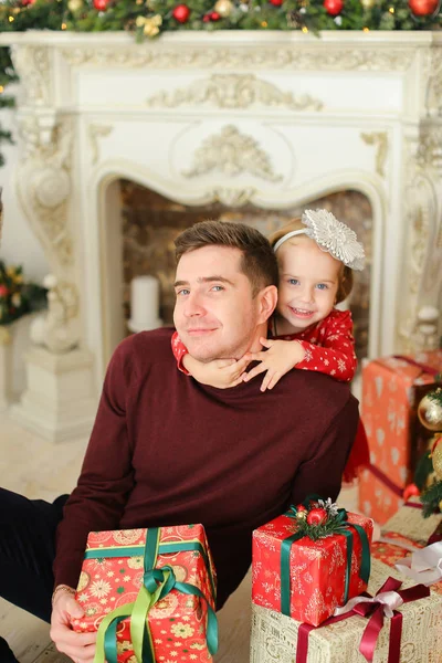 Pai sentado com a pequena filha perto de lareira decorada e mantendo presentes . — Fotografia de Stock