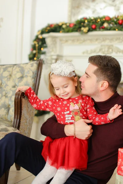 Feliz padre sentado con su hijita cerca de la chimenea decorada y manteniendo regalos . — Foto de Stock