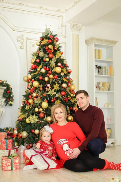 Niña sentada con el padre y la madre embarazada cerca del árbol de Navidad y guardando regalos . — Foto de Stock