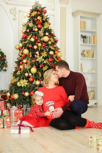 Pequeña hija europea sentada con el padre y la madre embarazada cerca del árbol de Navidad y guardando regalos . — Foto de Stock