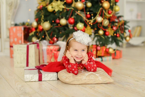 Petite fille blonde portant une robe rouge couchée près du sapin de Noël et des cadeaux . — Photo