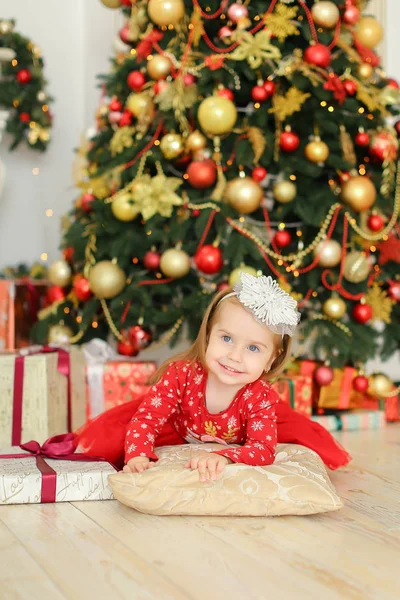 Kleines hübsches Mädchen in rotem Kleid liegt neben Weihnachtsbaum und Geschenken. — Stockfoto