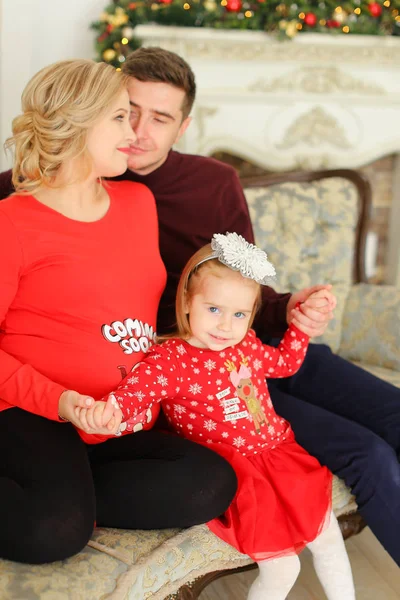 Criança feminina vestindo vestido vermelho sentado com pai e mãe grávida perto de lareira decoreated . — Fotografia de Stock