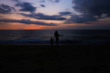 Mutlu anne ile küçük kız deniz kıyısında gündoğumu arka planda çalışan siyah silüeti.