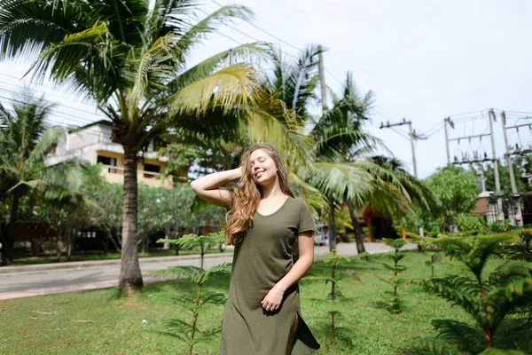 Jovem mulher feliz vestindo vestido cáqui e de pé perto de palmas no fundo . — Fotografia de Stock