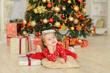 Küçük mutlu kız giyiyor kırmızı elbise Noel ağacının yanında yalan ve sunar.