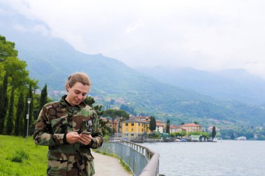 Kamuflaj ceketi yanında tırabzan, Como Gölü ve Alpler Dağı arka planda smartphone chat yapan giyen genç beyaz turist. Akdeniz ve Turizm, modern teknoloji kavramı.