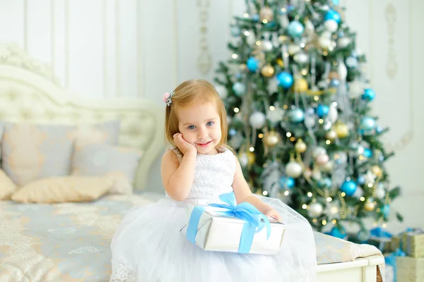 Kleines kaukasisches Mädchen in weißem Kleid hält Geschenk in der Nähe des geschmückten Weihnachtsbaums im Schlafzimmer. — Stockfoto