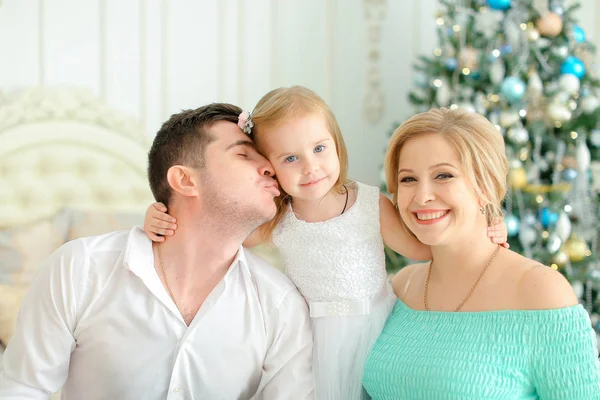Pequena filha sentada com beijando pai e mãe loira perto da árvore de Natal decorada . — Fotografia de Stock