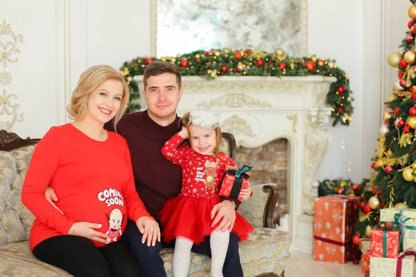 Mujer rubia embarazada sentada con el hombre y la hija cerca del árbol de Navidad y chimenea decorada . — Foto de Stock
