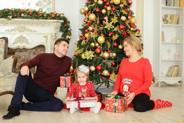 Kleine hübsche Tochter sitzt mit Vater und schwangerer Mutter am Weihnachtsbaum und bewahrt Geschenke auf. — Stockfoto