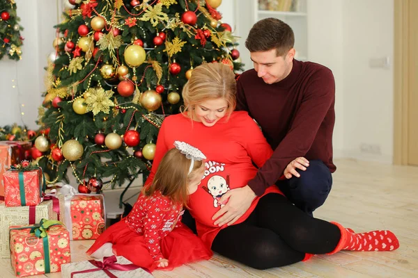 Mulher grávida europeia sentada com o marido abraçando barriga e filha perto de presentes sob a árvore Chistma . — Fotografia de Stock