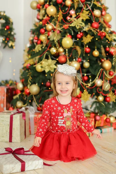 Kleines lächelndes weibliches Kind in rotem Kleid sitzt auf dem Boden in der Nähe von Geschenken und Weihnachtsbaum. — Stockfoto