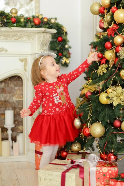 Kleines Mädchen in rotem Kleid schmückt Weihnachtsbaum am Kamin und Geschenke. — Stockfoto