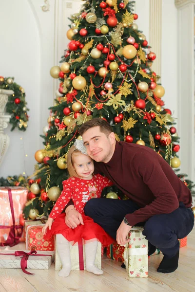 Pequeña hija bonita con vestido rojo abrazando padre cerca del árbol de Navidad y regalos . — Foto de Stock