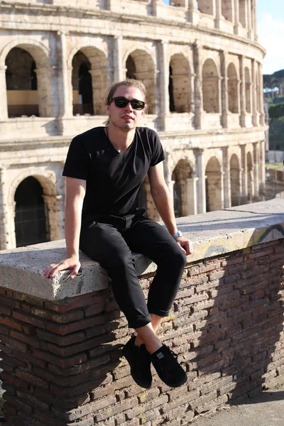 Joven turista caucásico en gafas sentado en la pared de ladrillo en el Coliseo fondo en Roma, Italia . — Foto de Stock