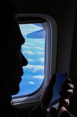 Uçak penceresinde yakınındaki smartphone kullanan genç adam silüeti portre.