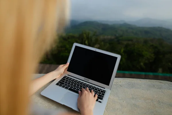 Chica rubia joven que trabaja con el ordenador portátil en el fondo de las montañas verdes, con camisa gris . — Foto de Stock