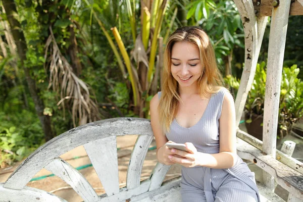 Jonge blonde vrouw hatting door smartphone met palmen in achtergrond, zittend op de schommel. — Stockfoto